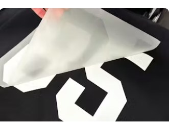 Transfer application sample from the original website; illustrates carrier film and artwork edges, not a verified GMB customer job. Carrier film peeling from a white number transfer on black fabric