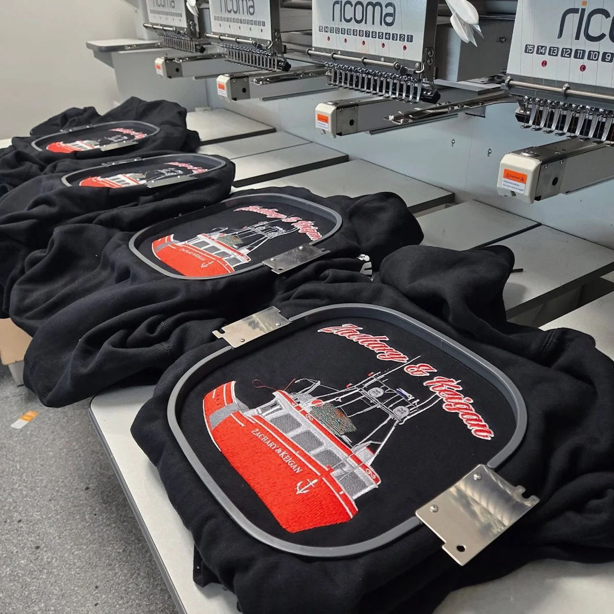 Photo from GMB's existing apparel gallery; shown for decoration and product planning, not current stock availability. Black garments held in embroidery hoops beneath machine needles for logo stitching